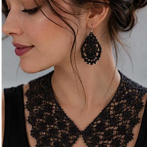 May include: A black lace collar necklace featuring an intricate floral pattern and a matching pair of teardrop-shaped black lace earrings with a delicate filigree design, worn over a simple black sleeveless top with a rounded neckline.