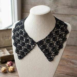 May include: A black crochet collar, displayed on a mannequin. The collar has an openwork design with a scalloped edge. The intricate, lace-like pattern is visible against the neutral background of the mannequin. The collar is a handmade item.
