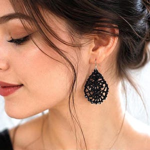 Pode incluir: Um brinco em forma de lágrima feito de fio preto com um intrincado padrão de renda vazada. O brinco apresenta um design delicado e pendente com um gancho de cor prateada, exibindo uma textura tecida detalhada em toda a peça.