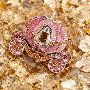 May include: A pink and gold pumpkin carriage brooch with sparkling crystals. The carriage has intricate details, including a door with a bear-shaped window. The wheels and accents are in shades of pink and orange, set against a gold background.