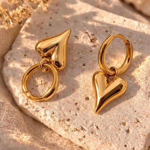 Könnte beinhalten: Ein Paar goldfarbene herzförmige Ohrringe. Ein Ohrring hat einen Herzanhänger, der an einem Reifen befestigt ist, der andere einen Herz-Charm, der an einem Reifen hängt. Die Ohrringe werden auf einer strukturierten, hellen Oberfläche präsentiert.