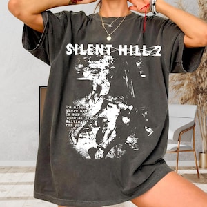 May include: Dark gray t-shirt with the text "SILENT HILL 2" in white block letters. Below the text is a white graphic and the words "I'm alone there now In our 'special place' Waiting for you" in a white font. The shirt has short sleeves.