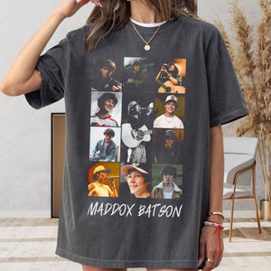 May include: A charcoal gray short-sleeve t-shirt featuring a collage of nine photographic images of a person in various settings, including playing guitar and wearing hats. The text MADDOX BATSON is printed in white in a stylized font at the bottom.