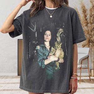 Könnte beinhalten: Ein anthrazitfarbenes Kurzarm-T-Shirt mit einem Grafikdruck einer Person in einem grünen Overall, die eine rote Katze hält. Der Druck zeigt eine Szene aus dem Film Alien mit der Figur Ellen Ripley und der Katze Jonesy in einer dunklen Umgebung.