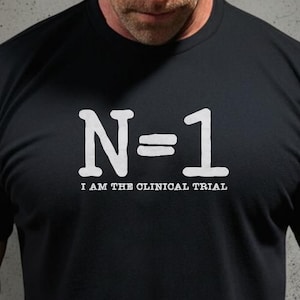 以下が含まれることがあります： 胸の中央に白い文字でN=1とプリントされた黒い半袖Tシャツ。N=1の下には、より小さな白いフォントでI AM THE CLINICAL TRIALというテキストが記されています。Tシャツはクルーネックで、標準的なフィット感のデザインです。