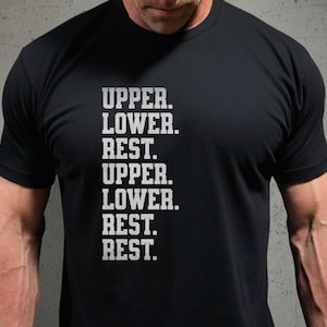 Può includere: T-shirt nero con testo bianco. Il testo recita "UPPER. LOWER. REST. UPPER. LOWER. REST. REST. REST." La maglietta è uno stile girocollo e sembra essere fatta di un materiale morbido. La maglietta è probabilmente per gli appassionati di fitness.