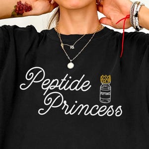 May include: Black t-shirt with the words "Peptide Princess" in a white script font. A small illustration of a vial with a crown is next to the text. The shirt is a casual style.