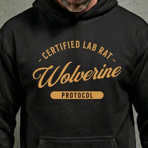 以下が含まれることがあります： 黒色のパーカー。金色の文字で「- CERTIFIED LAB RAT - Wolverine PROTOCOL」と書かれています。パーカーには、ドローストリングフードとフロントポケットが付いています。