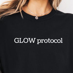 以下が含まれることがあります： 黒いクルーネックTシャツ。白い文字で「GLOW protocol」とプリントされています。柔らかい素材でできており、ゆったりとしたフィット感です。テキストは胸の中央に配置されています。