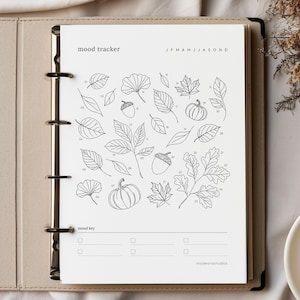Può includere: Una pagina di pianificazione per il monitoraggio dell'umore bianca con illustrazioni a linee nere di varie foglie, ghiande e zucche. La pagina presenta elementi numerati da 01 a 31, il testo mood tracker, J F M A M J J A S O N D, mood key e madeorastudios.