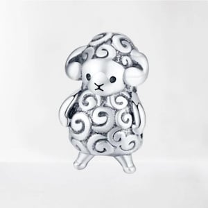 May include: A silver sheep charm with detailed curly wool patterns. The sheep has small, rounded ears and a face with a black 'X' mouth and two black dot eyes. The charm is standing on two small legs, and is a decorative item.