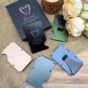 May include: A collection of metal wallets in various colors, including black, green, blue, and rose gold. Each wallet features a minimalist design with a money clip or stand. One black wallet is engraved with "Brent Groomsman 07/06/26" and a tuxedo graphic.