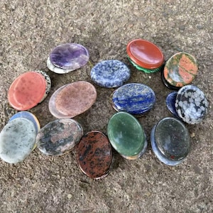 Könnte beinhalten: Eine Sammlung oval geformter, polierter Steine in verschiedenen Farben, darunter Lila, Rot, Blau, Grün und Braun. Die Steine sind auf einer rauen, grauen Oberfläche angeordnet und zeigen ihre natürlichen Texturen und Muster. Diese Steine eignen sich ideal für Meditation oder Dekoration.