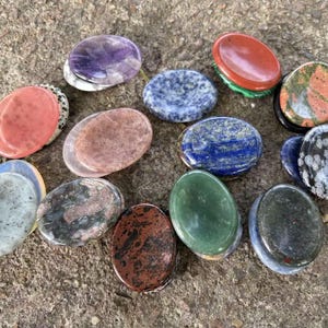Op de afbeelding: Een verzameling van vijftien gladde, ovaalvormige gepolijste stenen in diverse kleuren en patronen, waaronder paarse amethist, diepblauwe lapis lazuli, groene aventurijn, rode jaspis, gespikkelde zwart-witte obsidiaan en gevlekt graniet.