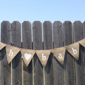 May include: A burlap banner with the words "oh baby" in white letters. The banner is strung on a wooden fence.