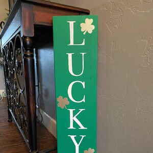 May include: A green wooden sign with the word "LUCKY" written in white letters. The sign is decorated with gold shamrocks and a rainbow over a pot of gold.