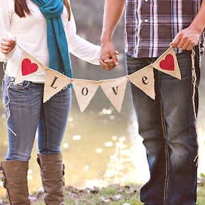 May include: A couple holding a burlap banner with red hearts and the word "LOVE" in black letters. They are standing in a grassy area with a body of water in the background.