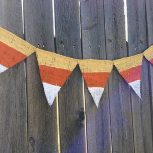 This picture is of a burlap banner with triangular shaped flags that look like candy corns. Each flag is hand painted with yellow on the top orange in the middle and white on the bottom.