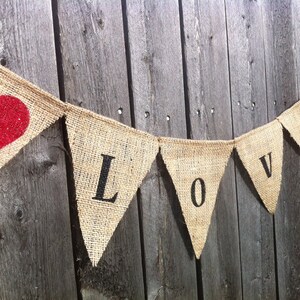 May include: A burlap banner with the word "LOVE" spelled out in black letters. Each letter is on a separate triangular flag. A red glitter heart is on the left side of the banner.