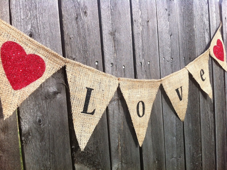 May include: A burlap banner with the word "LOVE" spelled out in black letters. Each letter is on a separate triangular flag. A red glitter heart is on the left side of the banner.