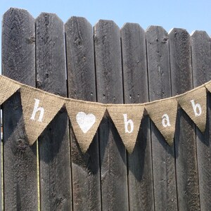May include: A burlap banner with white lettering that reads "Oh baby" with a white heart in between the words. The banner is hanging on a wooden fence.