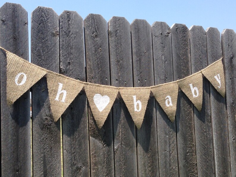 May include: A burlap banner with white lettering that reads "Oh baby" with a white heart in between the words. The banner is hanging on a wooden fence.