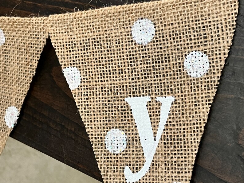 This photo shows an adorable gender neutral triangular flag baby banner perfect for a baby shower. The letters are hand painted in white. There are white polka dots with iridescent glitter paint on every flag.