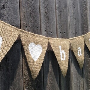 May include: A burlap banner with the word "baby" spelled out in white letters. The banner is strung on a wooden fence and has a white heart in the center.
