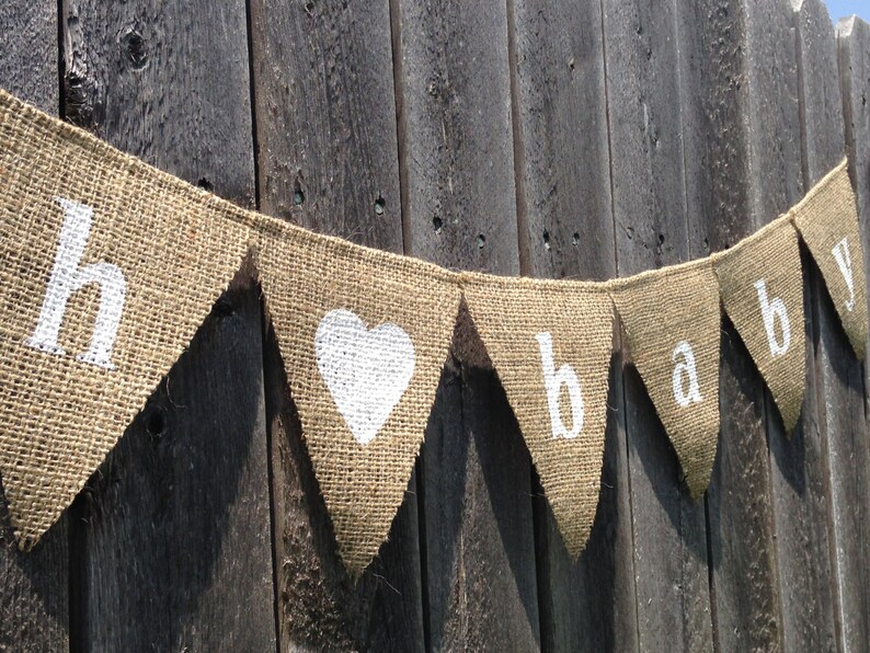 May include: A burlap banner with the word "baby" spelled out in white letters. The banner is strung on a wooden fence and has a white heart in the center.