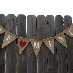 May include: A burlap banner with the words "BE MINE" in white letters and a red heart in the center. The banner is strung on a wooden fence.