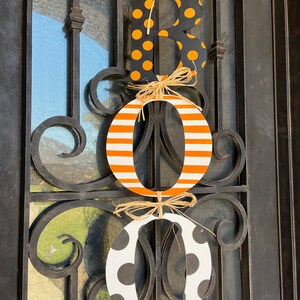 This is 3 letters hanging vertically saying boo for Halloween. The letters are tied together with raffia. The B is painted black with small orange polka dots, O is white with orange stripes, O with white with large black polka dots.