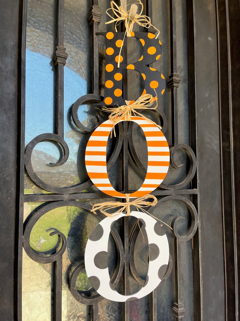 This is 3 letters hanging vertically saying boo for Halloween. The letters are tied together with raffia. The B is painted black with small orange polka dots, O is white with orange stripes, O with white with large black polka dots.