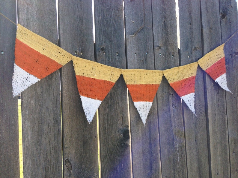 This picture is of a burlap banner with triangular shaped flags that look like candy corns. Each flag is hand painted with yellow on the top orange in the middle and white on the bottom.