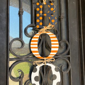 This is 3 letters hanging vertically saying boo for Halloween. The letters are tied together with raffia. The B is painted black with small orange polka dots, O is white with orange stripes, O with white with large black polka dots.