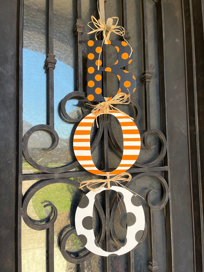 This is 3 letters hanging vertically saying boo for Halloween. The letters are tied together with raffia. The B is painted black with small orange polka dots, O is white with orange stripes, O with white with large black polka dots.