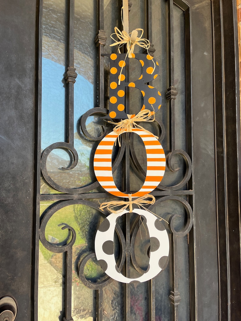 This is 3 letters hanging vertically saying boo for Halloween. The letters are tied together with raffia. The B is painted black with small orange polka dots, O is white with orange stripes, O with white with large black polka dots.