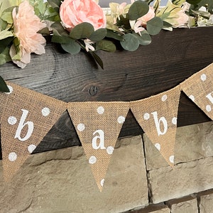 This photo shows an adorable gender neutral triangular flag baby banner perfect for a baby shower. The letters are hand painted in white. There are white polka dots with iridescent glitter paint on every flag.