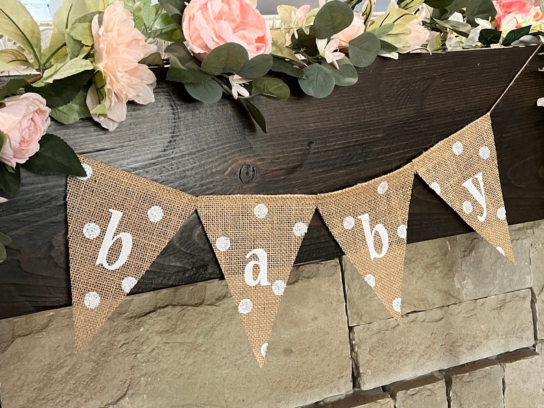 This photo shows an adorable gender neutral triangular flag baby banner perfect for a baby shower. The letters are hand painted in white. There are white polka dots with iridescent glitter paint on every flag.