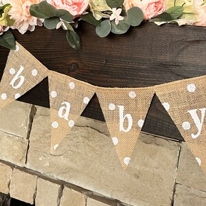 This photo shows an adorable gender neutral triangular flag baby banner perfect for a baby shower. The letters are hand painted in white. There are white polka dots with iridescent glitter paint on every flag.