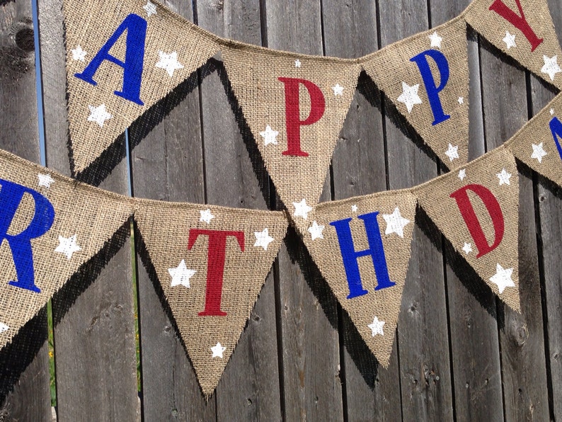This is a burlap banner that says happy birthday. The letters are hand painted in alternating blue and red letters. There are three to four white stars of different sizes on each flag.