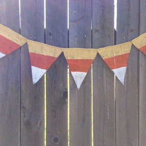 This picture is of a burlap banner with triangular shaped flags that look like candy corns. Each flag is hand painted with yellow on the top orange in the middle and white on the bottom.