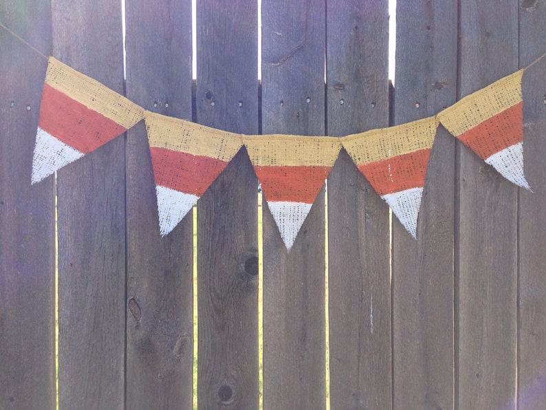 This picture is of a burlap banner with triangular shaped flags that look like candy corns. Each flag is hand painted with yellow on the top orange in the middle and white on the bottom.