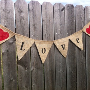 May include: A burlap banner with the word "LOVE" spelled out in black letters. The banner is strung between two wooden fence posts. There are two red hearts on the banner.