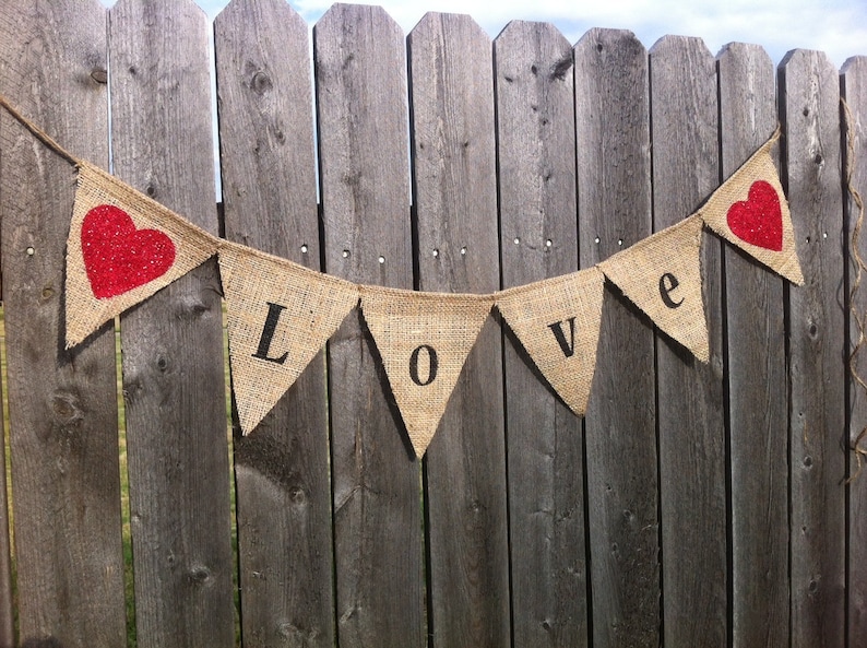 May include: A burlap banner with the word "LOVE" spelled out in black letters. The banner is strung between two wooden fence posts. There are two red hearts on the banner.