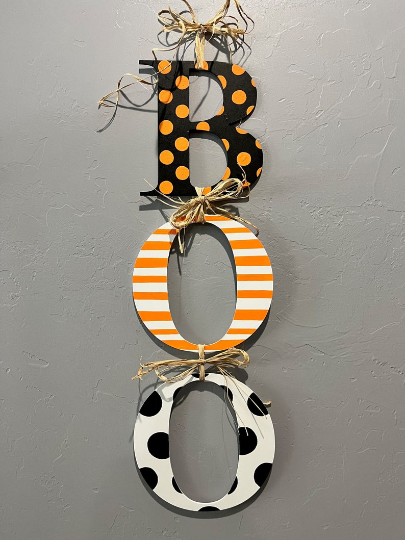 This is 3 letters hanging vertically saying boo for Halloween. The letters are tied together with raffia. The B is painted black with small orange polka dots, O is white with orange stripes, O with white with large black polka dots.