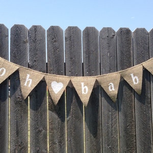 May include: A burlap banner with the words "Oh Baby" in white letters. The banner is strung on a wooden fence.