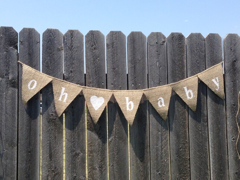 May include: A burlap banner with the words "Oh Baby" in white letters. The banner is strung on a wooden fence.
