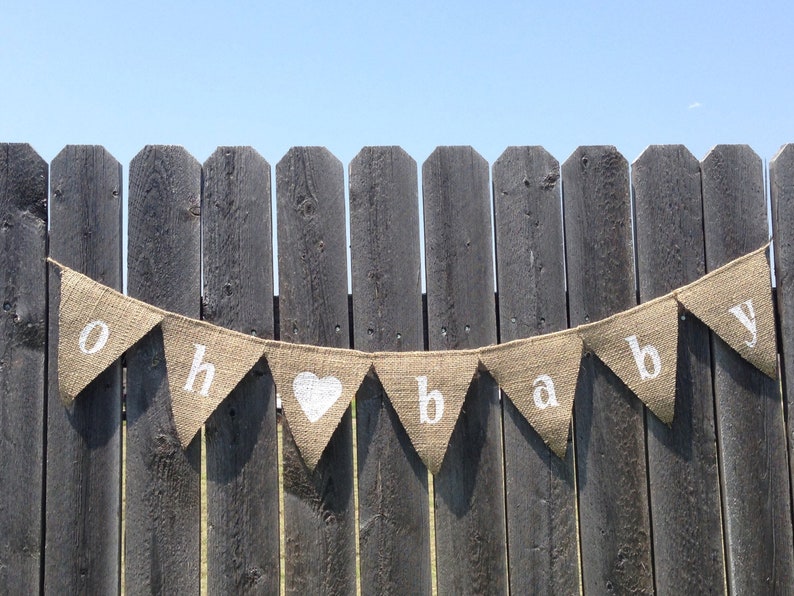 May include: A burlap banner with the words "oh baby" in white letters. The banner is strung on a wooden fence.