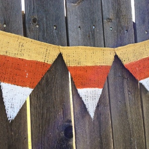 This picture is of a burlap banner with triangular shaped flags that look like candy corns. Each flag is hand painted with yellow on the top orange in the middle and white on the bottom.