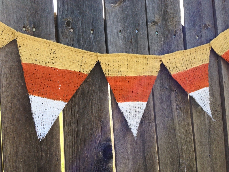 This picture is of a burlap banner with triangular shaped flags that look like candy corns. Each flag is hand painted with yellow on the top orange in the middle and white on the bottom.